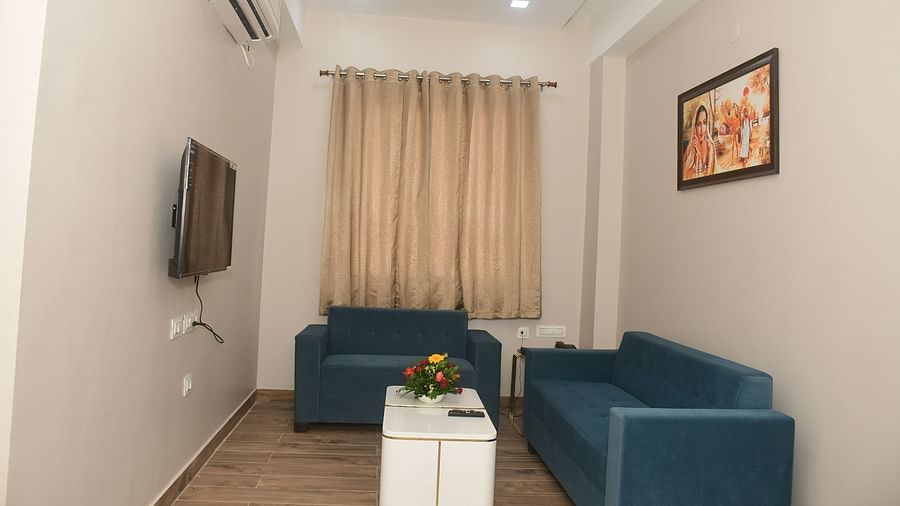A room features two blue sofas, a small white table, and a television against a white wall with a framed picture - Kankidham Lords Inn
