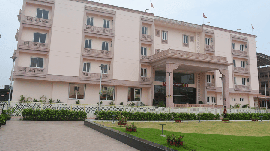 The front view of a modern, light-coloured hotel building with a covered entrance and green lawns in front is presented - Kankidham Lords Inn