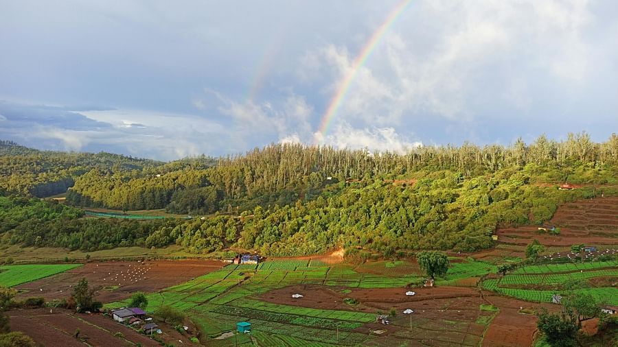 A vibrant rainbow arcs over a lush green valley with terraced fields and dense forests on the hillsides.