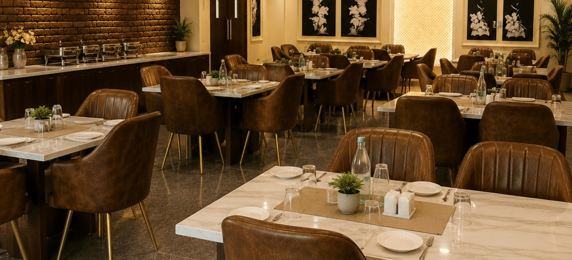 An indoor dining area or restaurant space with several set dining tables and brown chairs arranged under warm ceiling spotlights.