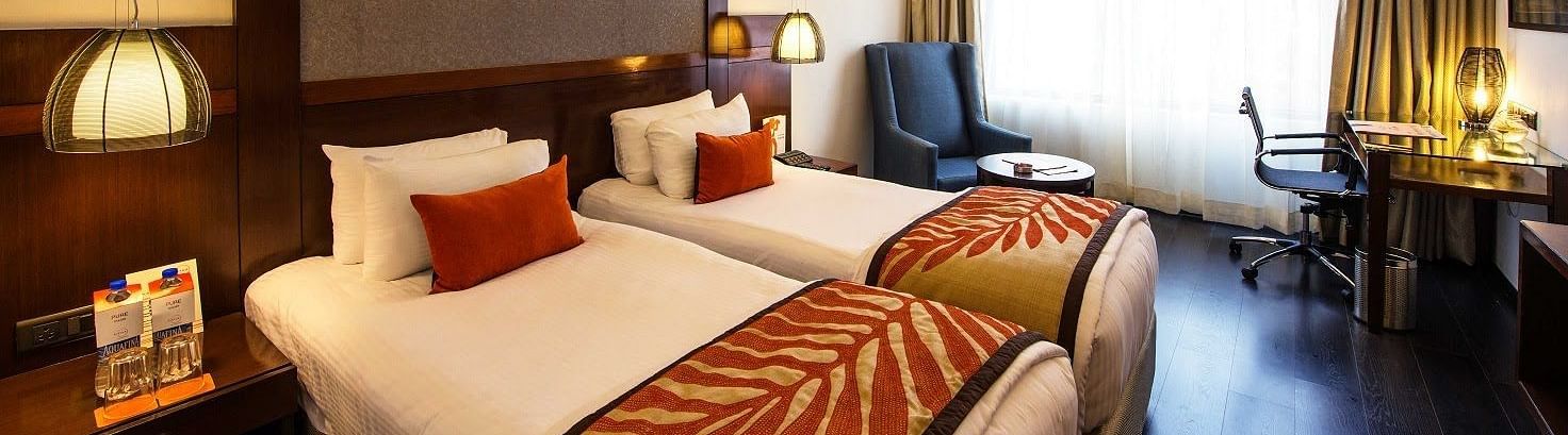 Deluxe Room at The Cunningham Hotel featuring twin beds with decorative runners, warm lighting, pendant lamps, a seating area, and a functional workspace near a large window with natural light