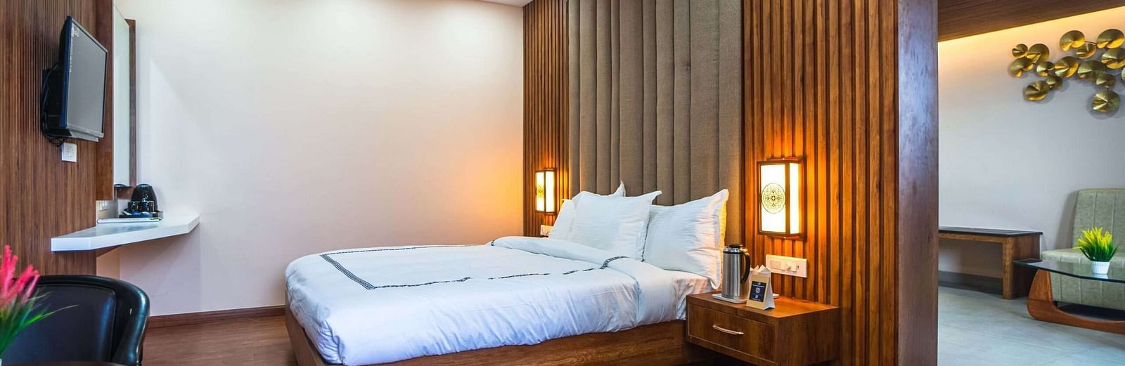 A hotel room at Mansha Palace features a bed, nightstands with lamps, a flat-screen TV, a ceiling fan, and a partial view into a seating area.