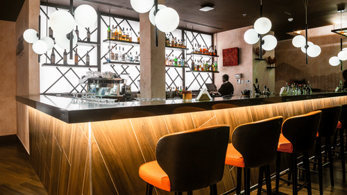 A bar area inside Asiatic Rooftop Bar and Restaurant, featuring stylish hanging lights, bar stools in orange and black, and a modern mirrored back bar.