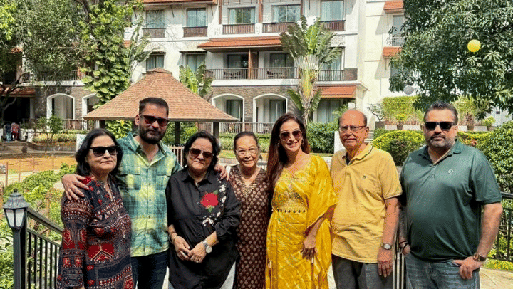A group of seven people, including two women in the center wearing sunglasses, posing for a photo outdoors in front of a resort building and a small gazebo.