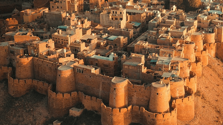 A high-angle aerial view of a dense, historic city with sandstone buildings and a large fort wall, all glowing in the light of the sun