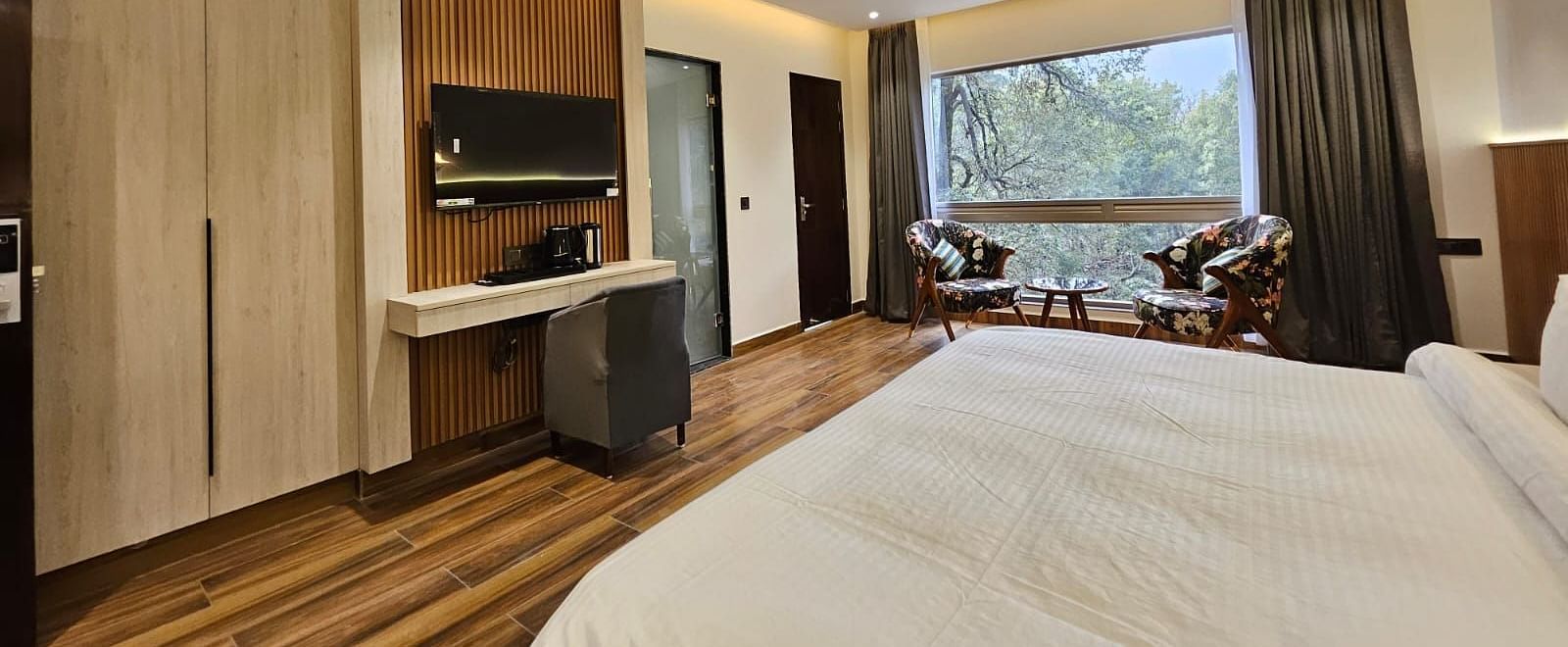A spacious premium room featuring the bed, flat screen TV, chairs, coffee table, work desk, and an expansive glass window overlooking the view - DLS The Emerald, Nainital
