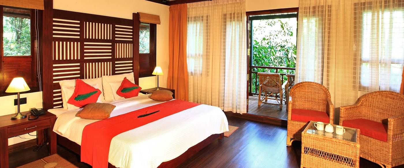 A bed placed in a room with two chairs and table, and a balcony that offers forest views | Vythiri Resort, Wayanad