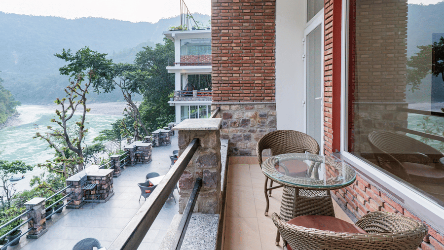 A balcony with a seating area featuring a round table and 2 bamboo woven chairs, overlooking some trees and a flowing river and mountains in the distance | Summit by the Ganges Beach Resort & Spa, Rishikesh