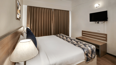Premium Studio at Starlit Suites Newtown, Kolkata features a double bed with a blue cushion and patterned throw, a wooden headboard, a bedside lamp, a wall-mounted TV, and a wooden bench.
