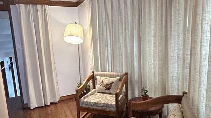 Sitting area in the Executive Suite of Cedar Inn, Darjeeling featuring two cushioned wooden chairs, a fridge, a lamp and curtained windows.