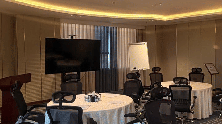 A conference room at Grand Iva Hotel, Abha featuring a round table with chairs, a television screen, and ceiling lights.