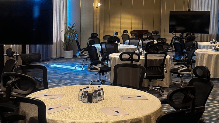 A meeting room at Grand Iva Hotel, Abha featuring a round table with office chairs, paper pads, and distant seating arrangements. 