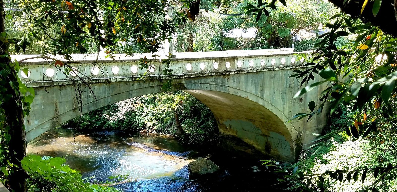 An arched bridge spans a narrow stream, framed by foliage in the foreground.