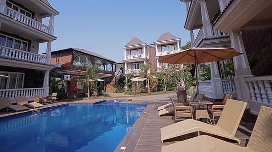 Poolside loungers facing elegant resort buildings under a clear sky - Grand Continent, Anjuna - A Sarovar Portico Affiliate Hotel 