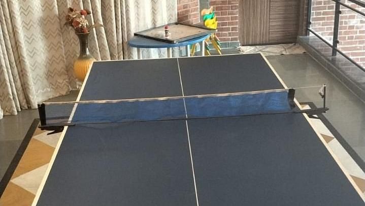 A table tennis setup indoors, featuring a blue table, a net, and paddles at Tisya Farms, Gurgaon.
