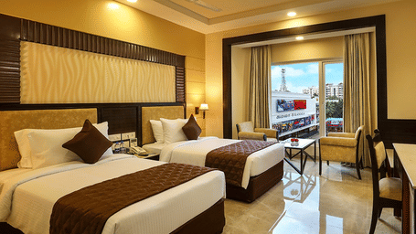 The room at Hotel Aaditya featuring a large bed, bedspreads, pillows, large windows covered with curtains, with a table, and a chair.