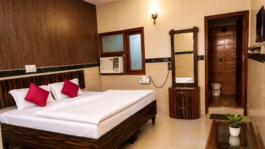 Bedroom interior of Hotel Goverdhan, Agra, featuring king size bed with white linen, 2 pillows, wooden headboard, window air conditioner, dressing table, mirror, table, and plant.
