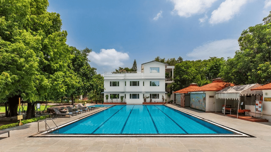A large outdoor swimming pool with lanes is situated next to a white building and green trees at Amantra Shilpi Resort, Udaipur.