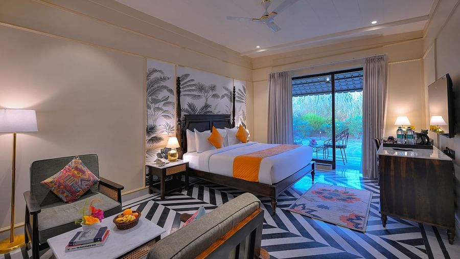 Superior Cottage featuring a four-poster bed with white linens, a colourful patterned runner and pillows, a patterned armchair, a wall-mounted TV, and sliding glass doors leading to a balcony - Singhvilas, Ranthambore