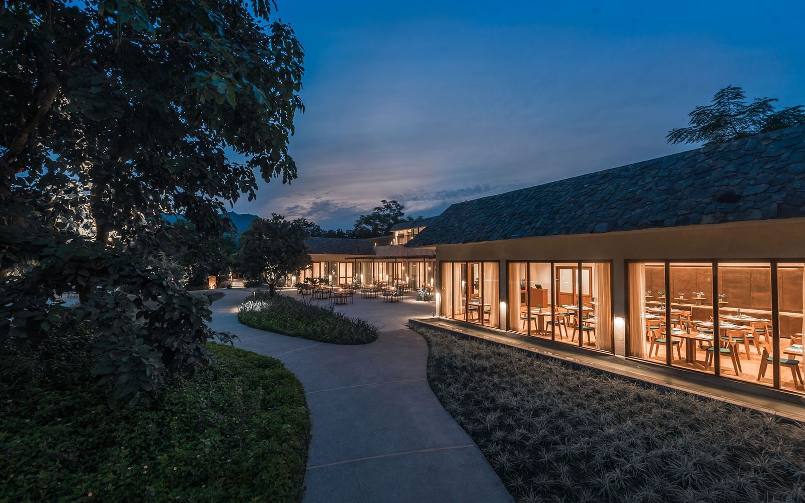 Exterior of a well-lit restaurant at Saraca  Resort & Spa Corbett with mountains in the backdrop during the blue hour