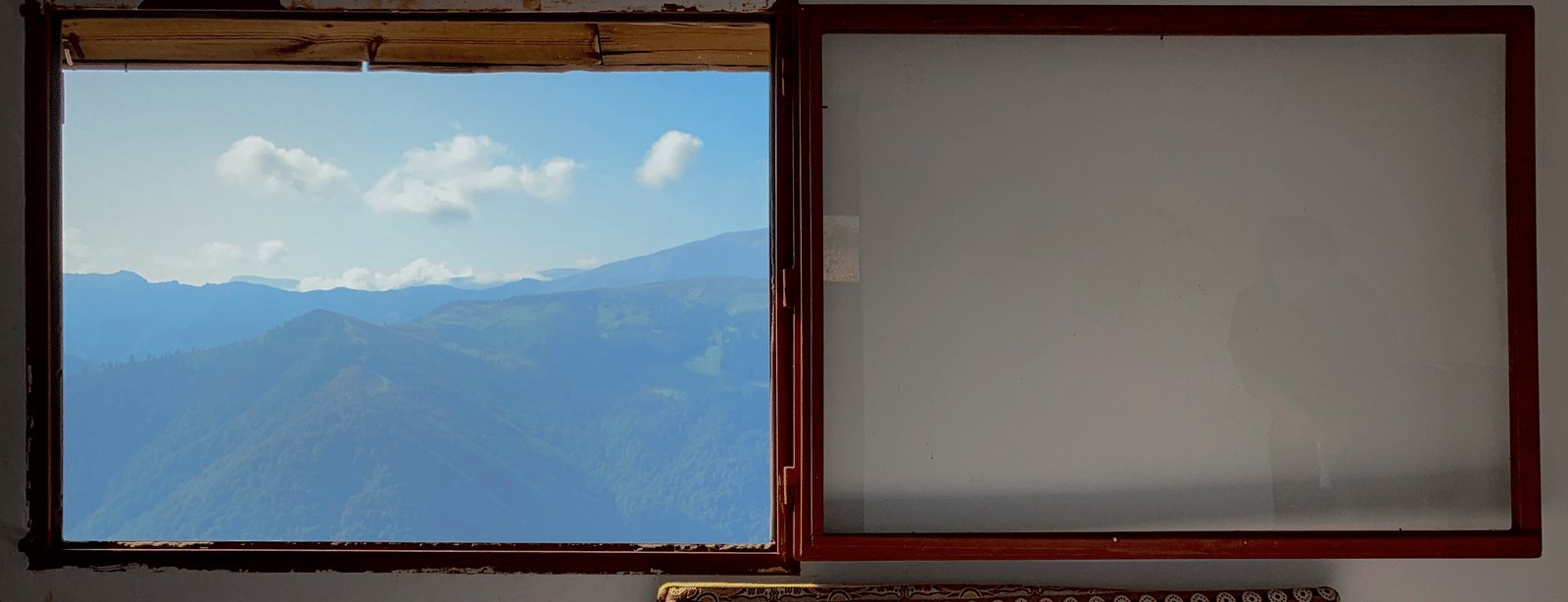 An interior wall with a view of green mountains and blue sky through an open window pane, next to a blank window and a rug below.