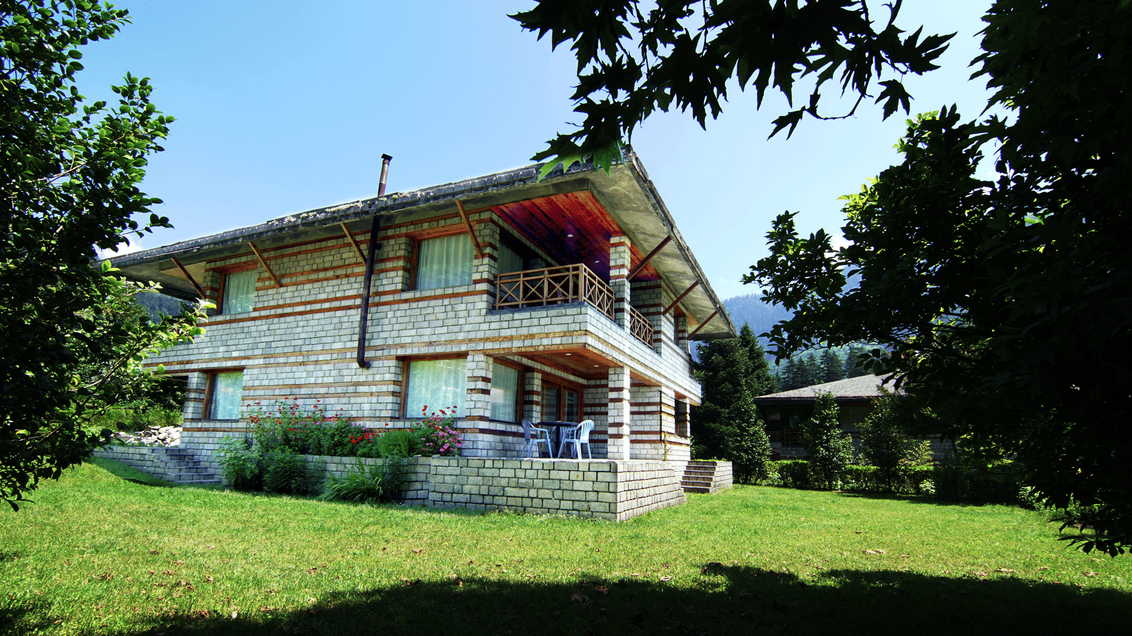 Exterior view of Lamrin Banon Resorts, Manali featuring a stone cottage, lush green lawn, and scenic mountain surroundings under a clear sky.