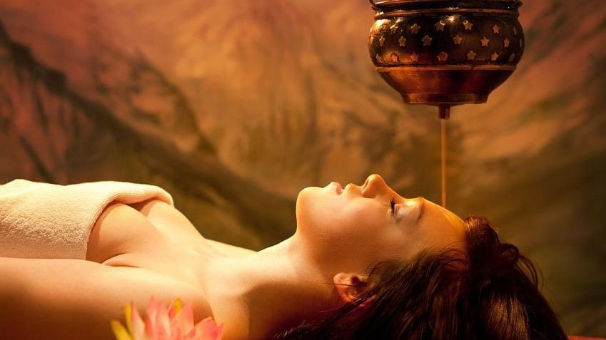 A serene woman is receiving an Ayurvedic Shirodhara treatment, with warm oil pouring from a brass pot onto her forehead.