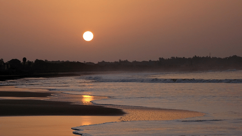 A sunset view over the sea with waves at the shore and mountains in the background.