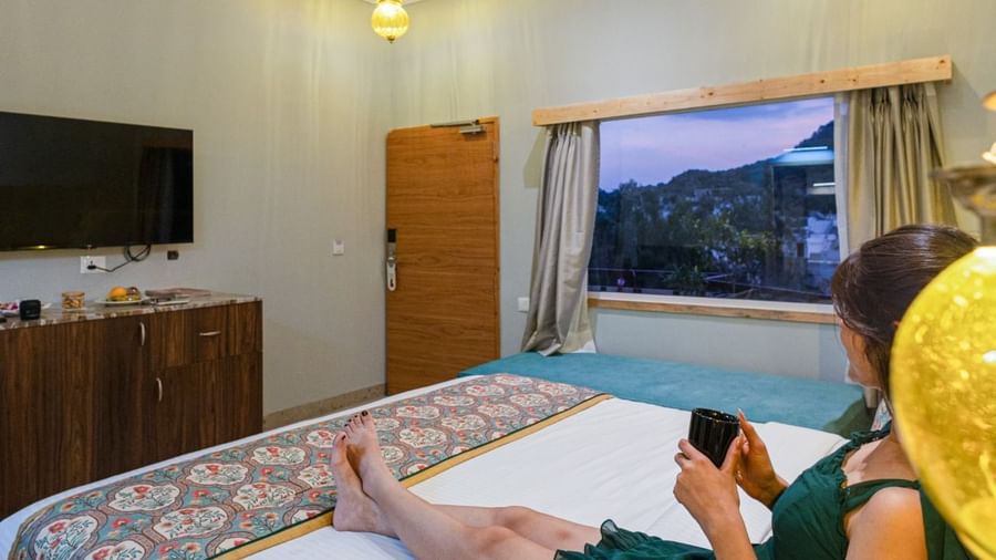 Guest relaxing on bed at Bamboo Saa Resort & Spa, Udaipur with hills view from window.