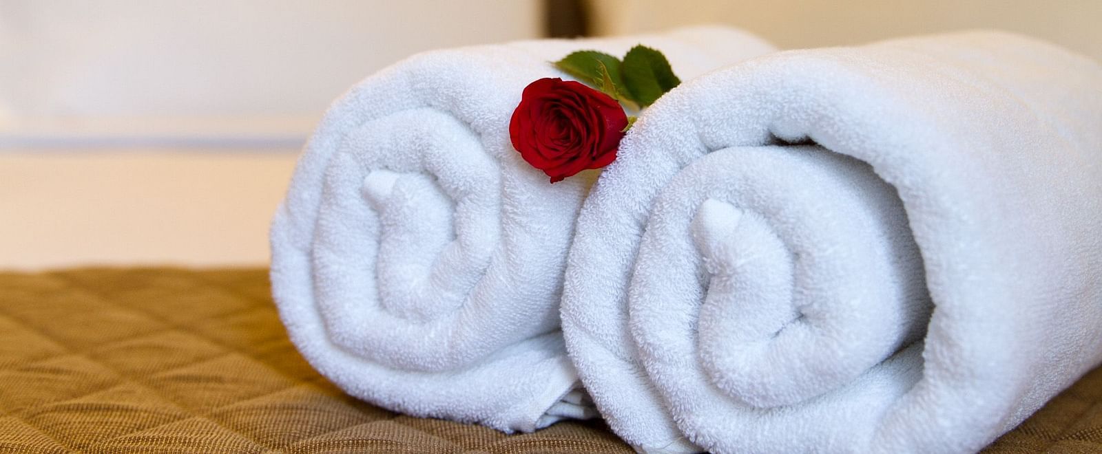 Two neatly rolled white towels are placed on a bed, adorned with a single red flower at Sanctum Suites Whitefield, Bangalore.