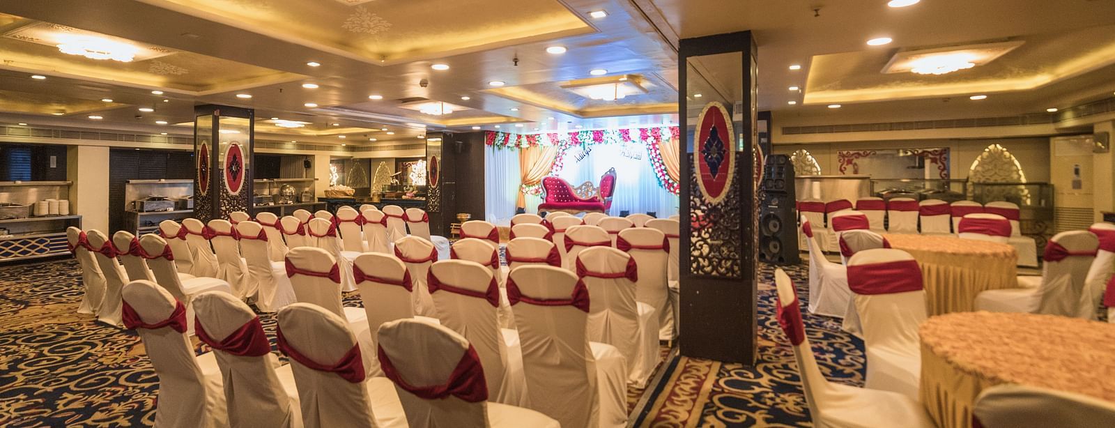 Maurya featuring rows of chairs with white covers and red sashes facing a stage area with draped fabric and lighting.