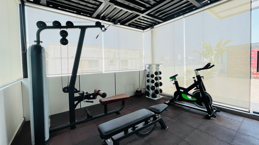 Rooftop gym at Hotel Kyzen Smart Stays Madhapur featuring a spinning cycle, weight rack, exercise bench, and glass panel walls with city views.