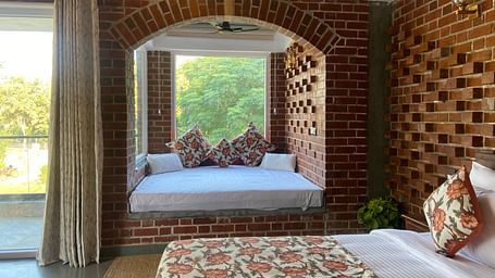 A bedroom at Tisya Farms, Gurgaon featuring a bed near a large arched window, brick walls, and a curtain on one side.