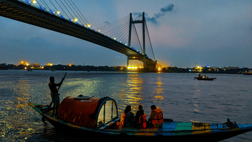 A boat with people on it travelling on a water body in front of the Howrah Bridge in Kolkata during twilight hour.
