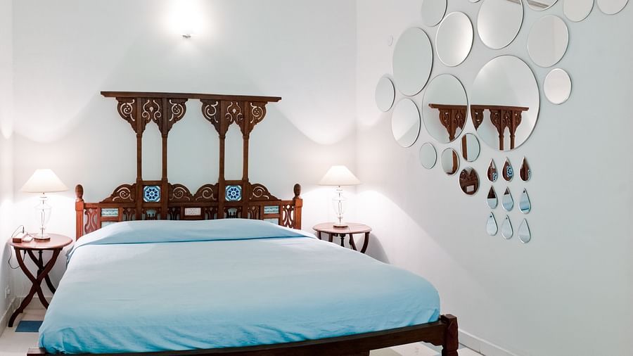 A bedroom with a wooden bed featuring a light-blue sheet, bedside tables with lamps, and a decorative arrangement of mirrors on the wall at Tijara Fort-Palace - 19th Century, Alwar.