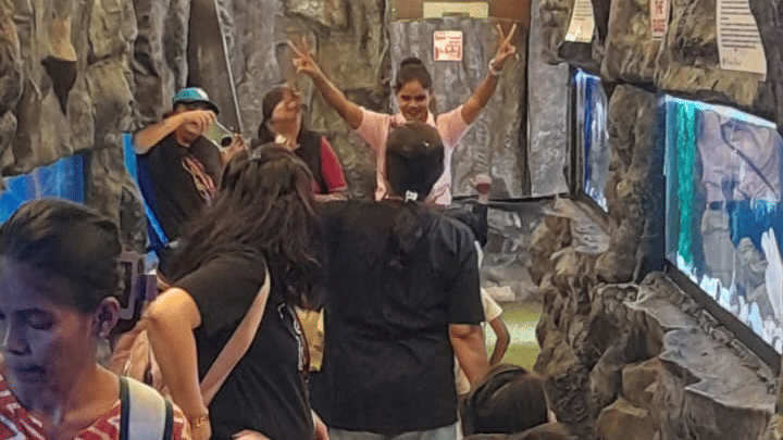 A vertical, brightly lit interior view of a crowded area at EsselWorld Fish Paradise, possibly a tunnel or hallway inside the aquarium, filled with many visitors.