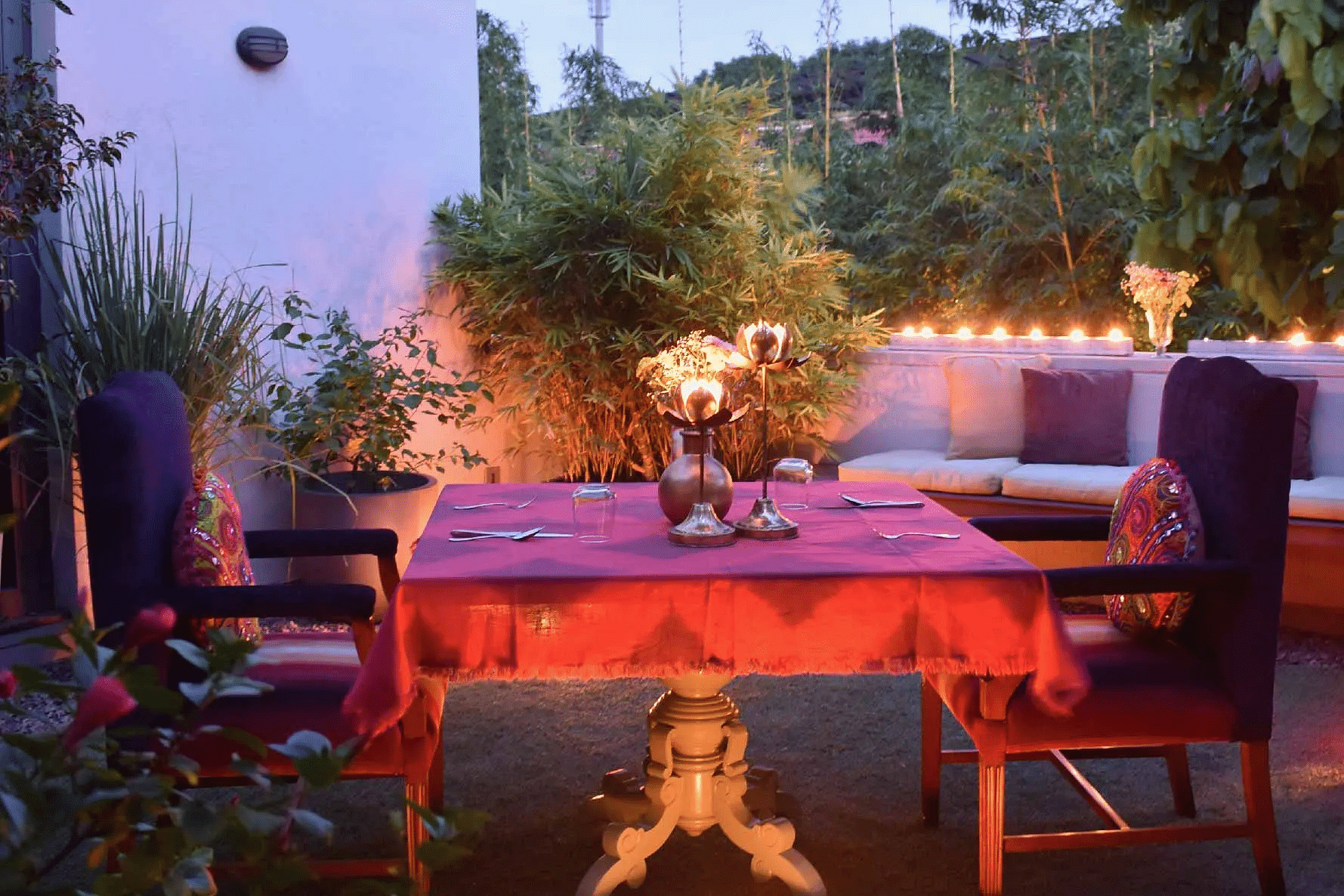 Evening patio setup with a red dining table, cushioned seating, soft lighting, and greenery all around at Karma Chalets.