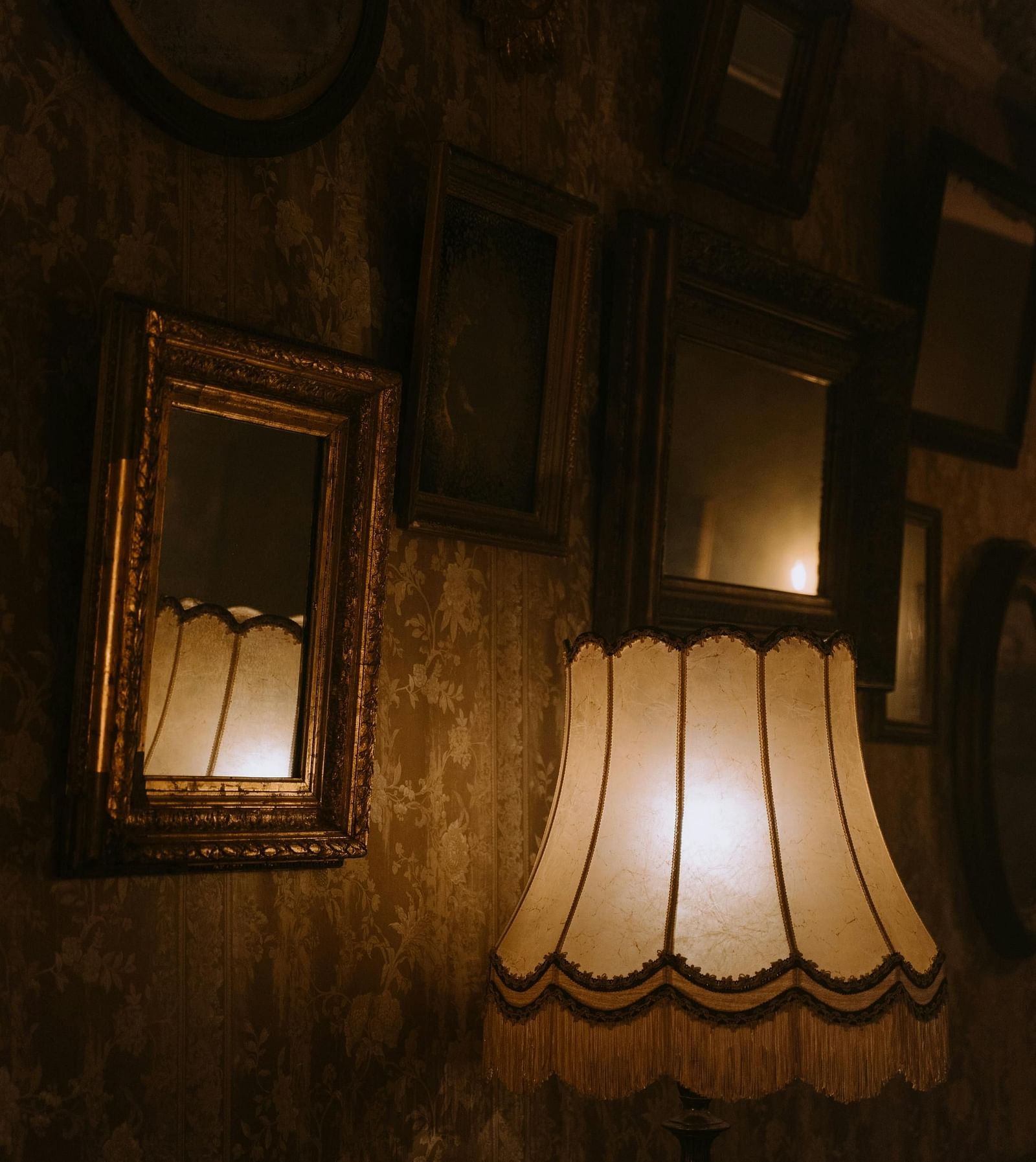 An old-school room with a desk lamp placed in front of a wallpapered wall with multiple frames.