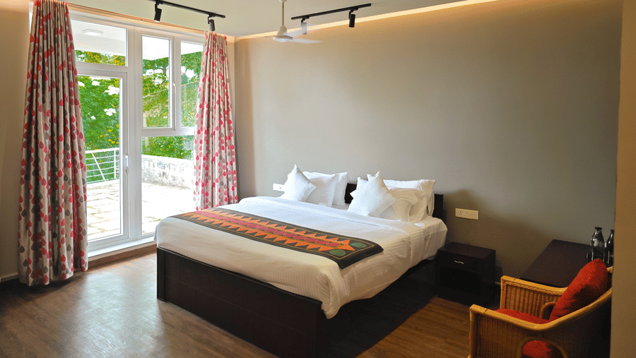 Bedroom in Starling room at Ibex Resorts, Coonoor (Tapas). A bed sits in the middle of the room with an armchair beside it. Curtains cover the windows in the background.