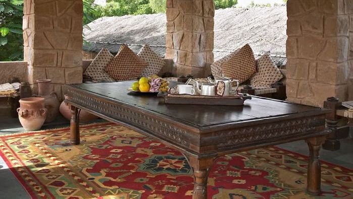 A dining area with open walls and a hut style roof featuring a huge table with food items, around it there are seating areas with cushion.