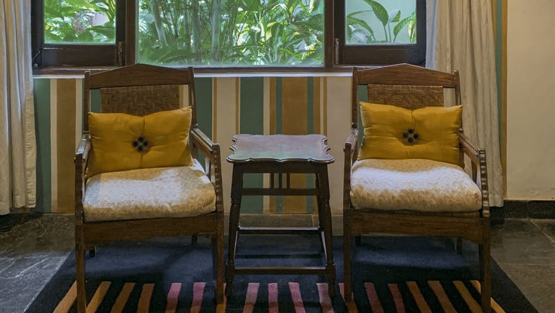 Window-side seating area with two cushioned chairs, a wooden table and a view of surrounding greenery.