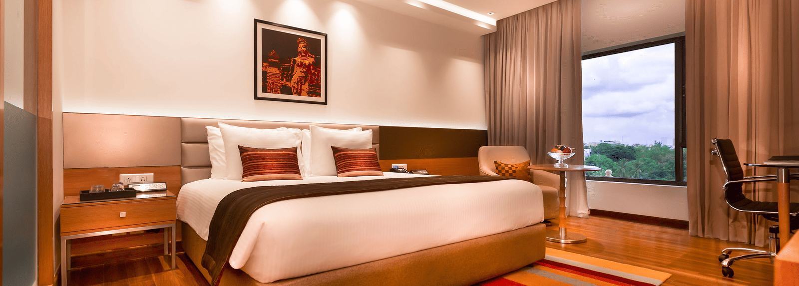 A modern hotel room featuring a king size bed, warm lighting, wooden flooring, a striped rug, and a large window with a view - Grand Chennai by GRT Hotels