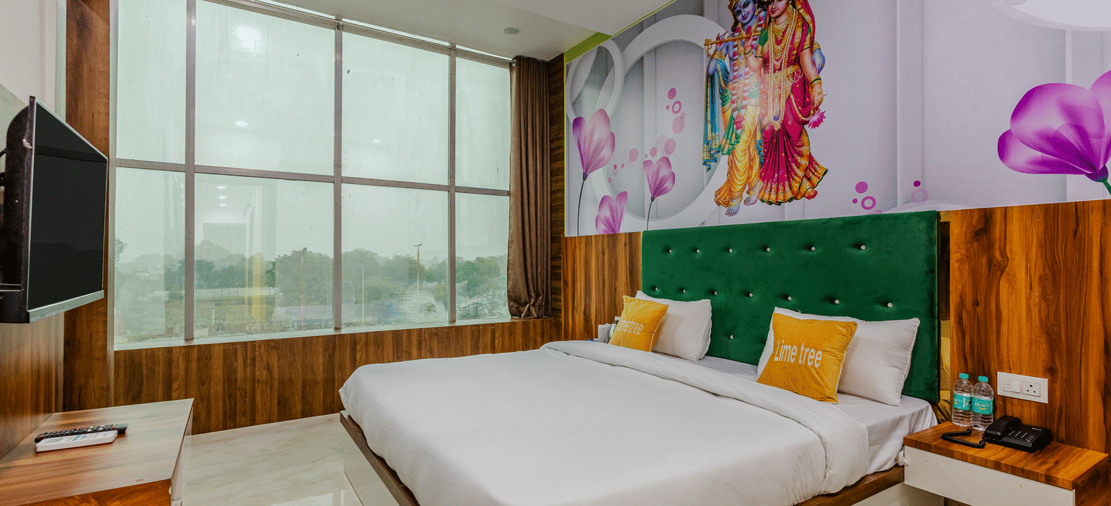 A bedroom with a green headboard, artwork above the bed, large windows, and a wall-mounted television - Lime Tree Hotels & Banquet, Vrindavan