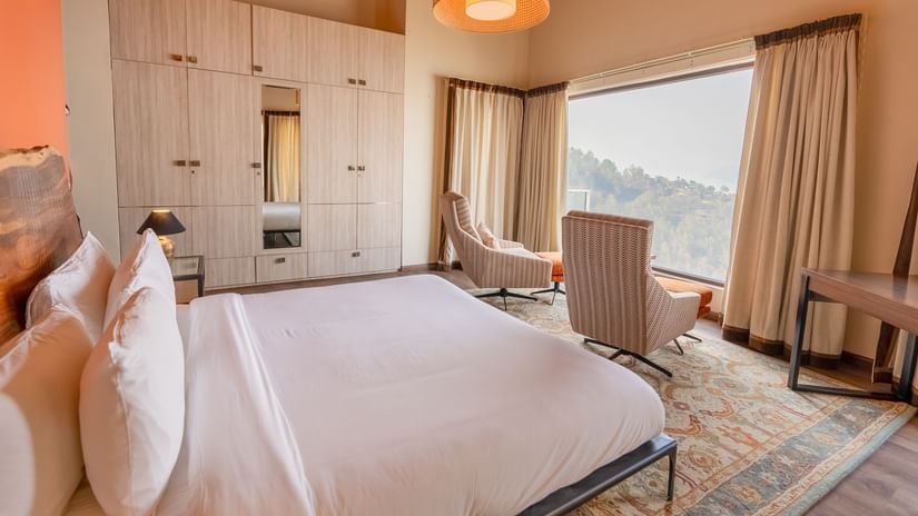 Bright, modern hotel room with a large bed, comfortable chair, and a big window offering scenic views of greenery and hills at  The Manor Luxury Apartments, Shimla.