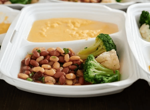 Multiple divided trays hold prepared meals with beans, broccoli, and a sauce, arranged on a surface.