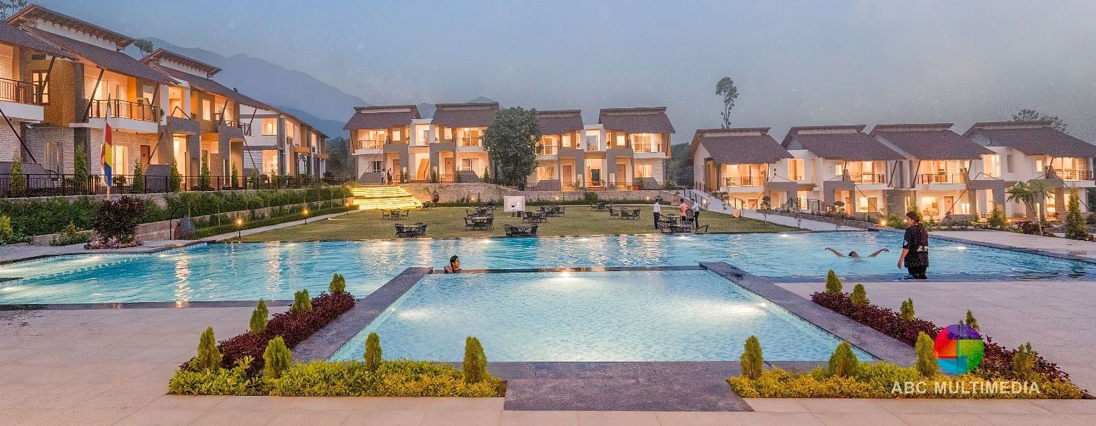 A large resort with multiple buildings surrounding a swimming pool, under a twilight sky.