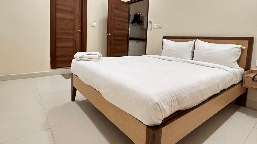 Modern bedroom at A M Suites with wooden bed frame, white bedding, folded towel, tiled floor, beige walls, air conditioning unit, and bright lighting creating a clean minimal look.