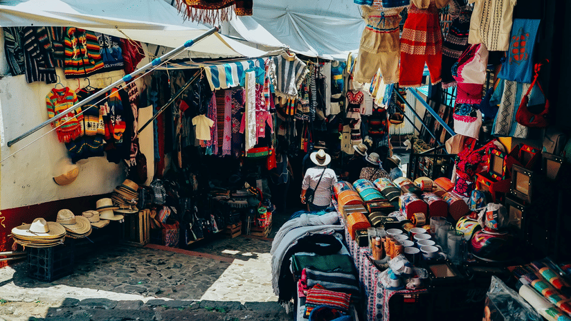 A street market with stalls selling clothes, bags, and textiles, with items displayed on tables and hanging from the stall frames.