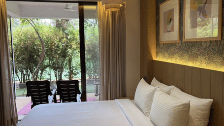 A king size bed with white linen faces a large glass sliding door revealing a balcony with 2 chairs and a garden view at Bougainvillea, Alibaug.