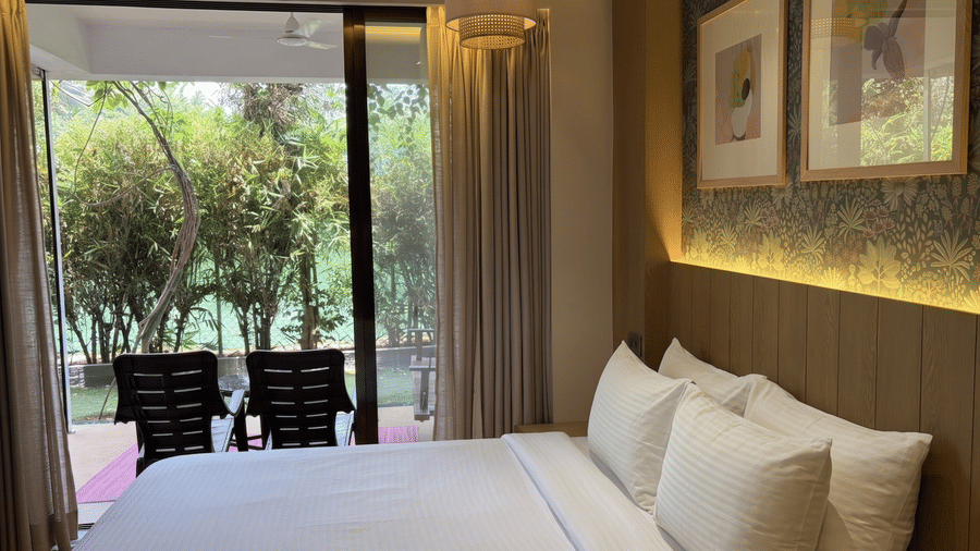 A king size bed with white linen faces a large glass sliding door revealing a balcony with 2 chairs and a garden view at Bougainvillea, Alibaug.
